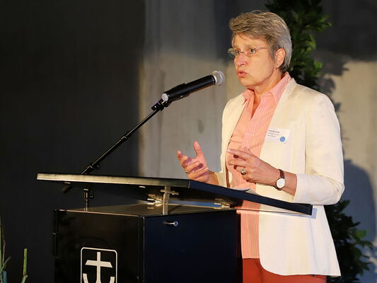 Eine Person spricht in ein Mikrofon hinter einem Rednerpult, dekoriert mit einem christlichen Symbol.