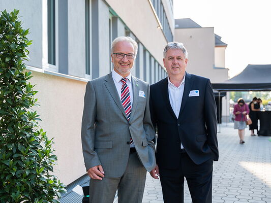 Zwei Männer in Anzügen vor einem Gebäude mit Pflanzen und Personen im Hintergrund auf einem Außenweg.