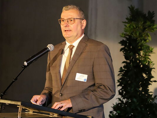 Ein Mann im Anzug steht an einem Rednerpult und spricht in ein Mikrofon, eine Pflanze im Hintergrund.
