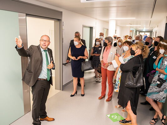 Eine Gruppe von Menschen in einem Flur, einige tragen Masken. Ein Mann zeigt etwas mit seiner Hand.