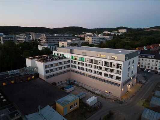 Ein modernes Gebäude mit mehreren Stockwerken und einer Dachterrasse bei Abenddämmerung in einer städtischen Umgebung.