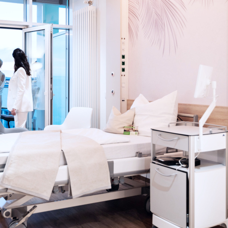 . Ein modernes Krankenhauszimmer mit Bett, medizinischem Wagen und zwei Personen, die am Fenster stehen.