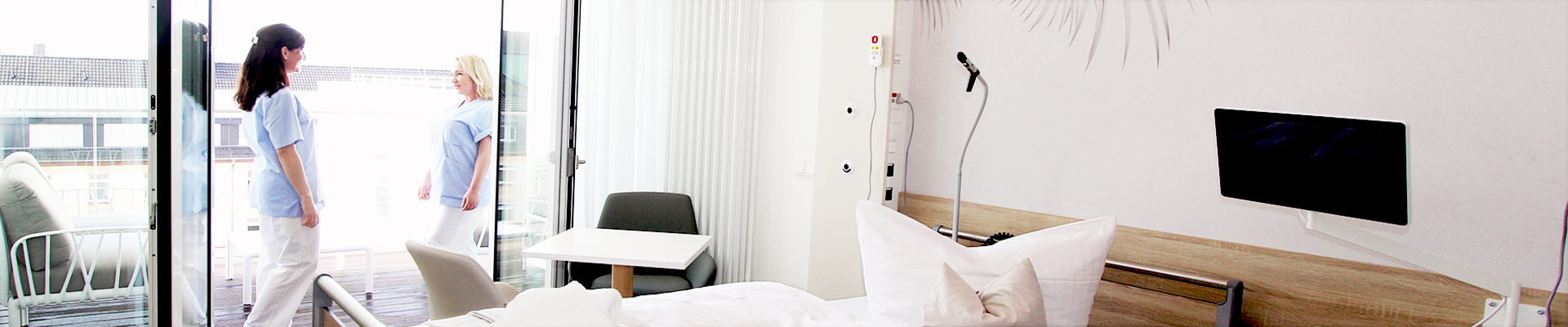 Ein Klinikzimmer mit Bett, Tisch, Stuhl, Terrasse; zwei Personen in blauem Kittel stehen draußen.