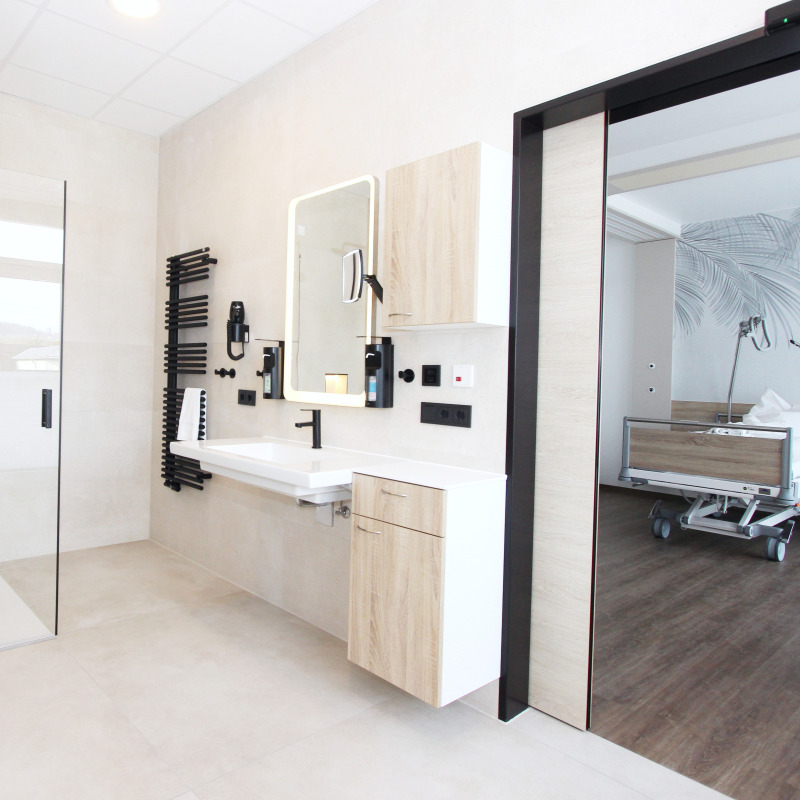 Ein modernes Badezimmer mit Waschbecken und Spiegel, daneben ein angrenzendes Zimmer mit Pflegebett.
