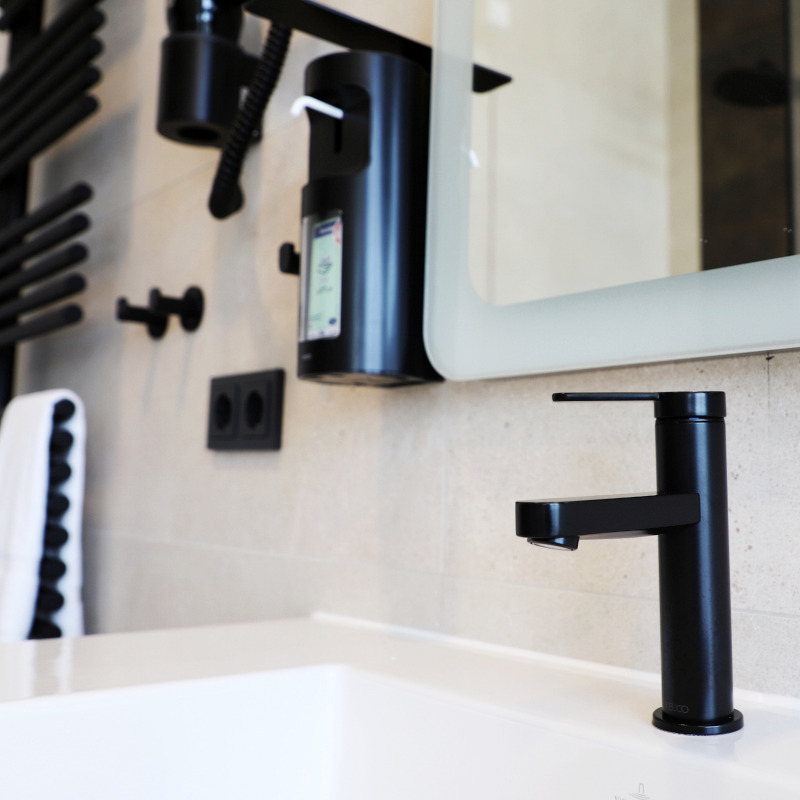 Modernes Badezimmer mit schwarzem Wasserhahn, Seifenspender und Handtuchhalter vor einem Spiegel.