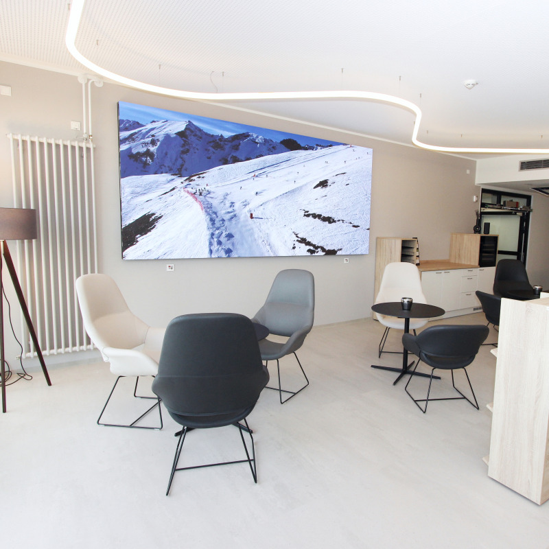 Ein moderner Warteraum mit Stühlen und Tischen, an der Wand ein großes Bild von schneebedeckten Bergen.