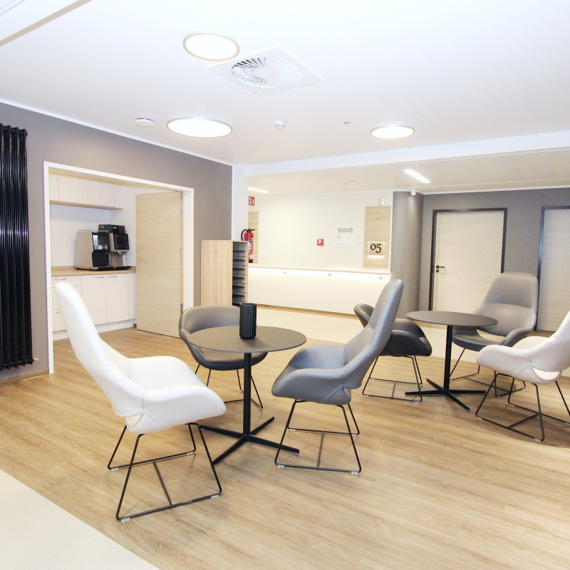 Moderner Warteraum mit Tisch, Stühlen, Kaffeemaschine und Holzboden in einem hellen, minimalistischen Design.