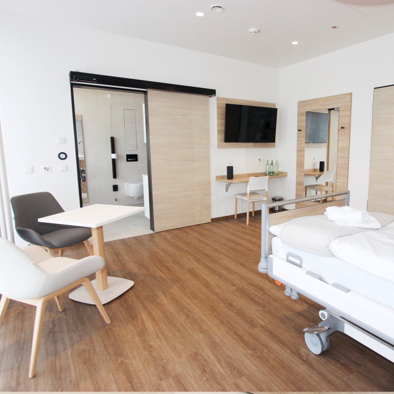Ein modernes Krankenhauszimmer mit Bett, Stühlen, Tisch, Fernseher und Badezimmer.