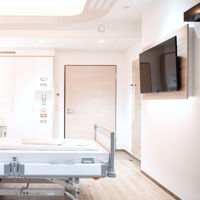 Ein modernes Krankenhauszimmer mit Bett, TV, Schränken und heller Beleuchtung.