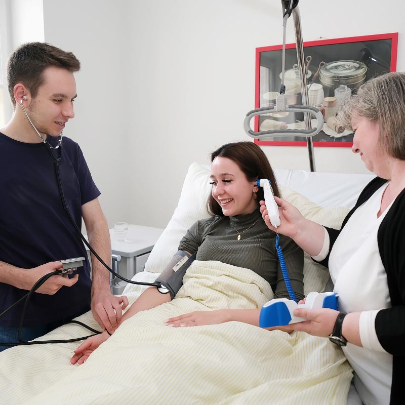 Eine Frau im Krankenhausbett wird von medizinischem Personal mit einem Blutdruckmesser und Thermometer untersucht.