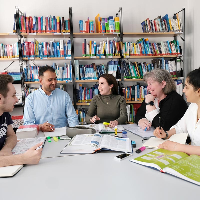 Fünf Personen studieren zusammen an einem Tisch, umgeben von Bücherregalen in einer Bibliothek.