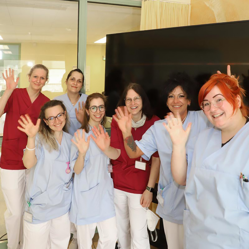 Eine Gruppe von Krankenschwestern und -pflegern in blauen und roten Kitteln lächelt und winkt freundlich.