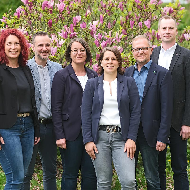 Eine Gruppe von sechs Personen steht lächelnd vor blühenden Sträuchern im Freien.