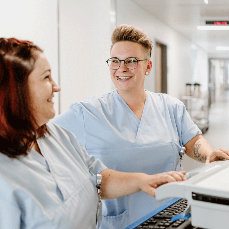Zwei Personen in blauer Arbeitskleidung lächelnd im Krankenhausflur, eine mit Brille und Tattoos.
