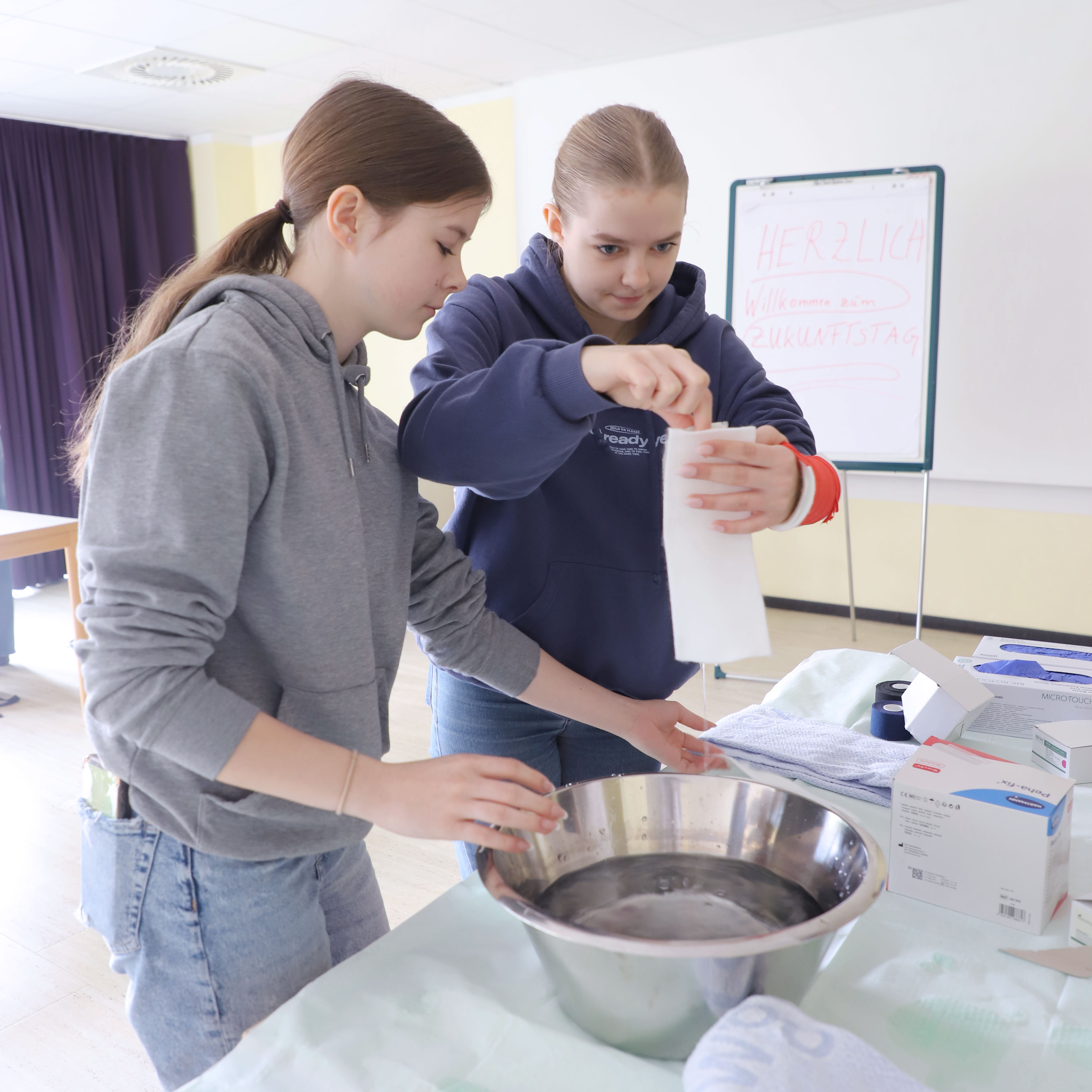. Zwei Mädchen, die an einem Tisch mit Wasser, Handtuchrollen und medizinischen Handschuhen arbeiten.