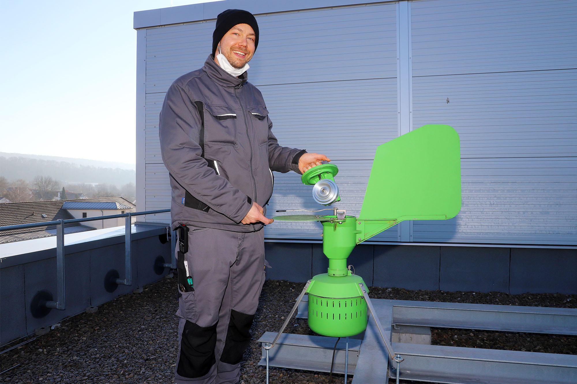Eine Person in einem Arbeitsanzug steht auf einem Dach neben einem großen grünen Gerät.