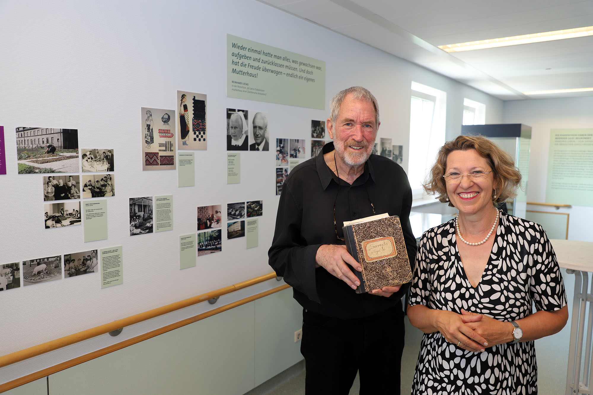 Zwei Personen posieren vor einer Wand mit Fotos und Text. Eine Person hält ein Notizbuch.