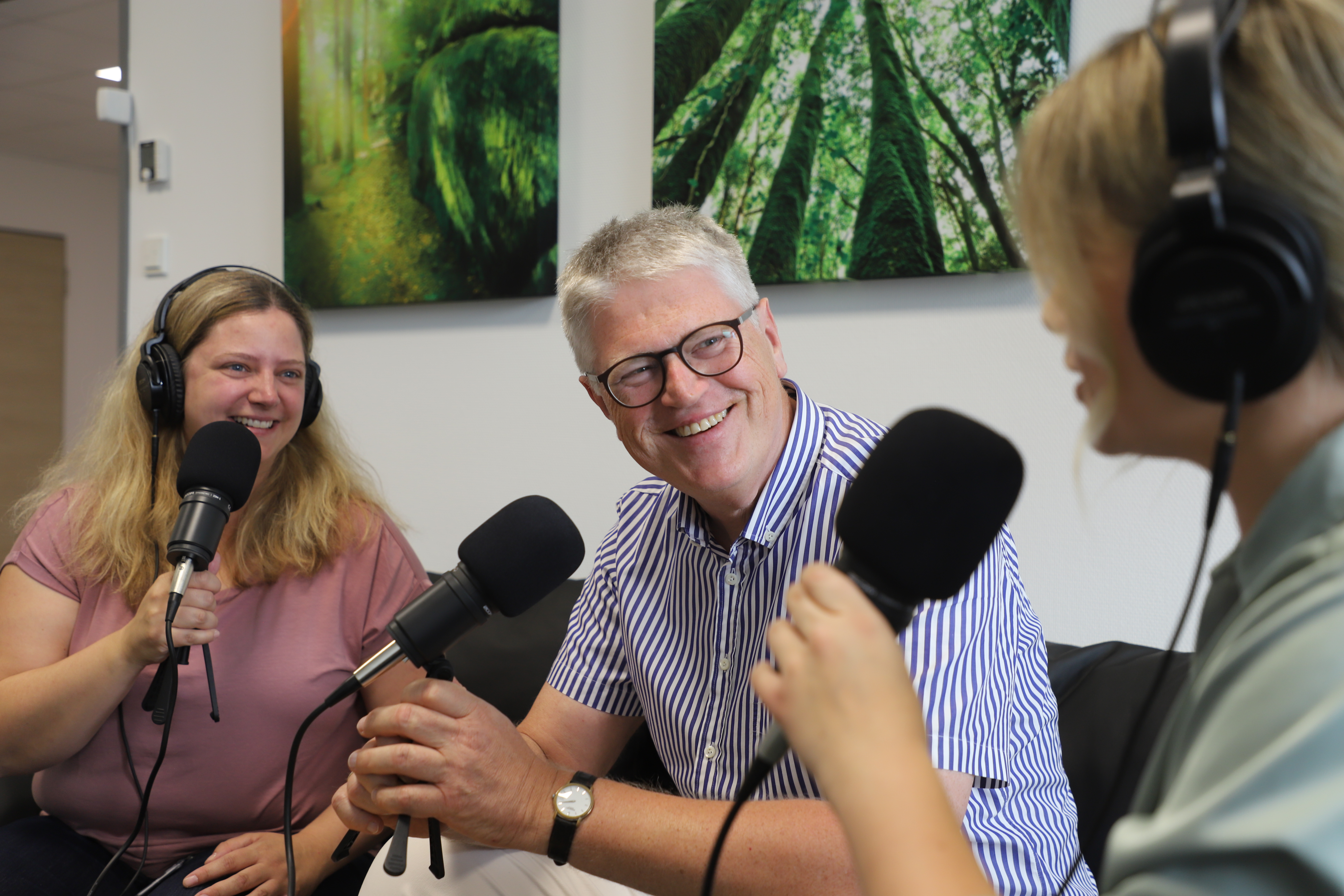 Drei Personen mit Mikrofonen und Headsets nehmen an einem Gespräch in einem Raum mit Waldkunstwerken teil.