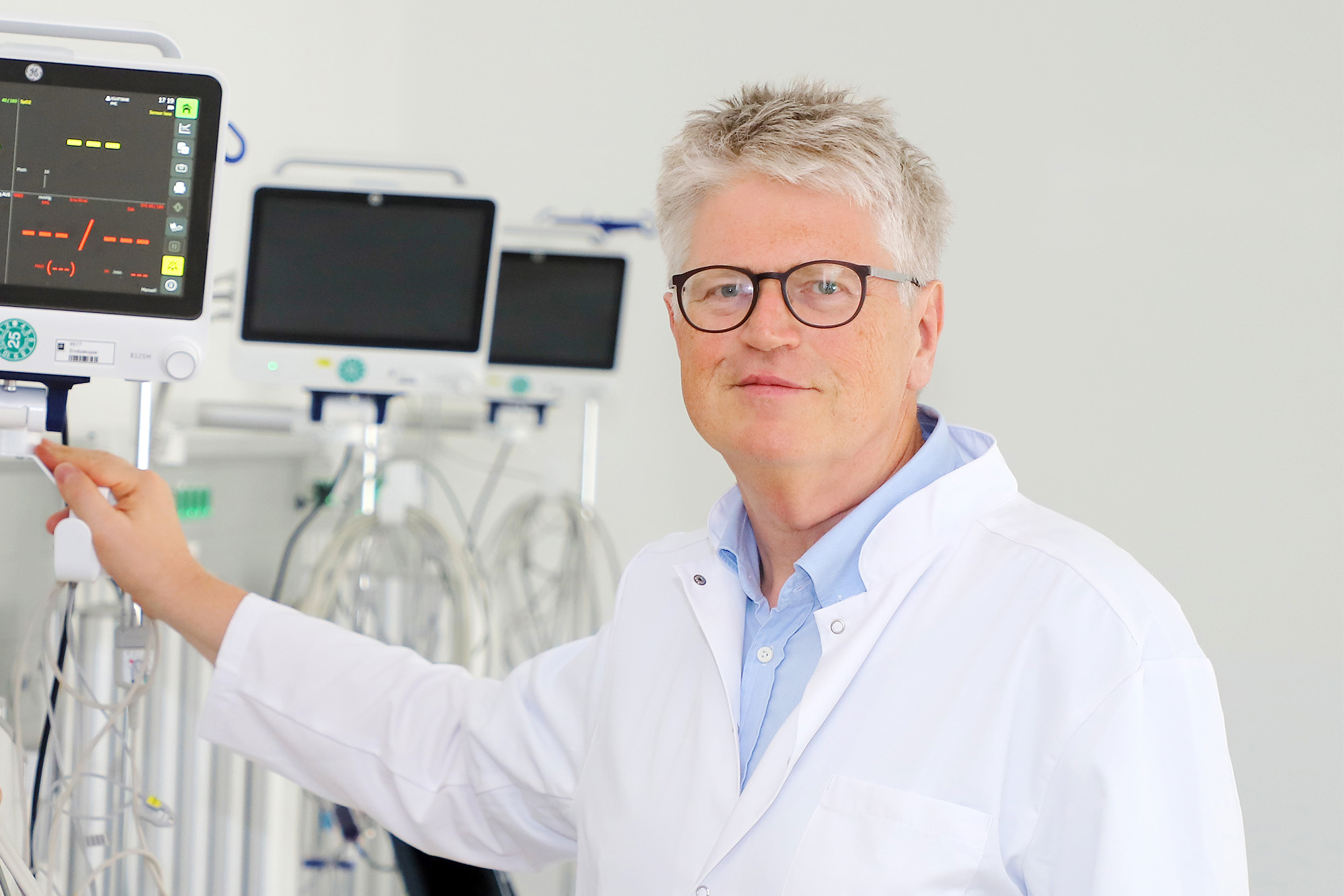 Ein Mann in einem weißen Kittel steht neben medizinischen Geräten, darunter ein Monitor mit Anzeigen.