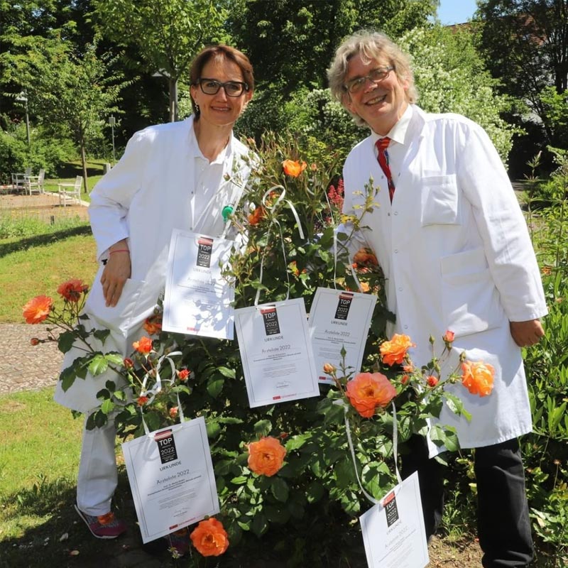 Zwei Personen in weißen Kitteln stehen bei einem Rosenstrauch mit Auszeichnungen daran.