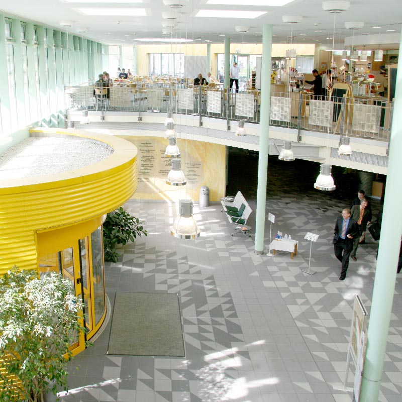 Ein heller Gebäudeeingang mit gelbem Eingang, Sitzbereich oben, und Menschen, die sich bewegen.