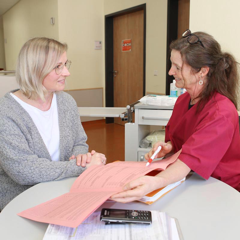 Zwei Frauen in einem Gespräch in einem Büro, mit einem rosa Formular und einem Stift auf dem Tisch.