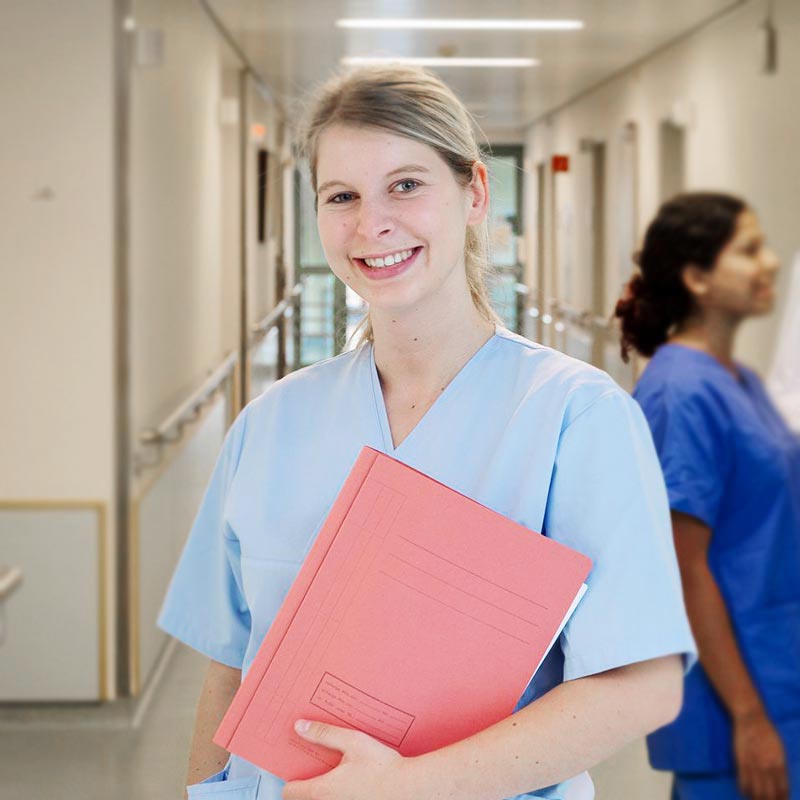 Eine Krankenschwester in blauer Kleidung hält eine rote Mappe in einem Krankenhausflur.