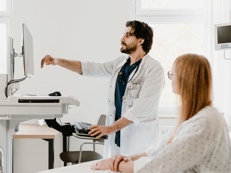 Ein Arzt zeigt einer Patientin auf einem Monitor etwas. Sie sind in einem Untersuchungsraum.