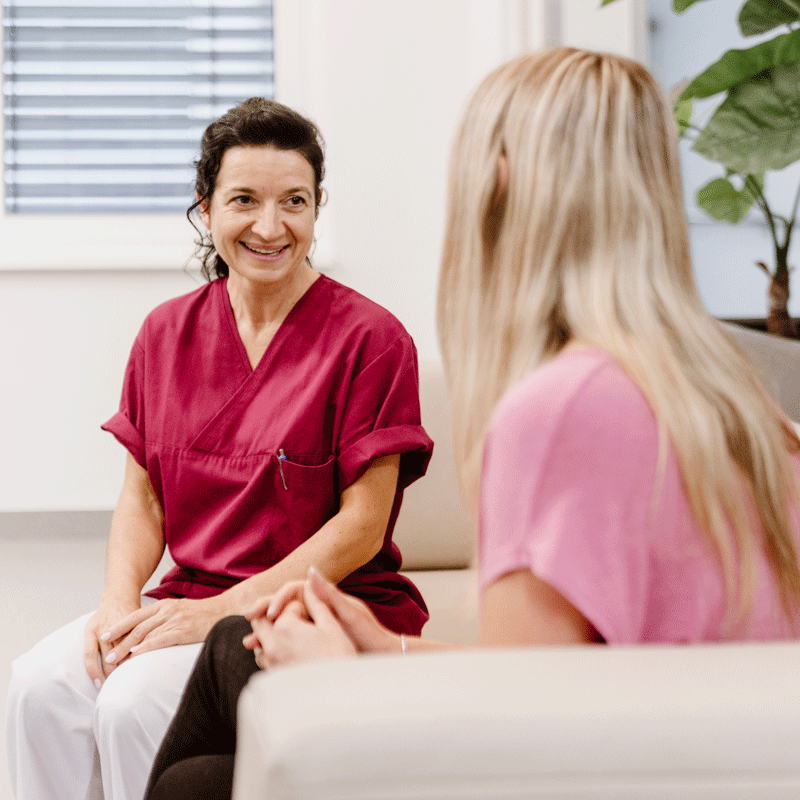 Zwei Frauen im Gespräch, eine in roter Arbeitskleidung, die andere in rosa, sitzen und lächeln in einem Büro.