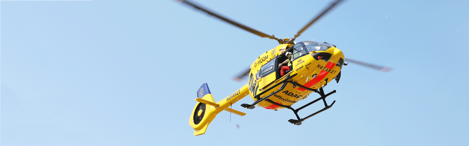 Ein gelber Rettungshubschrauber der ADAC Luftrettung fliegt mit offenem Seitentür gegen blauen Himmel.