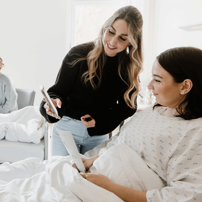 Eine Frau zeigt einer anderen Frau im Bett etwas auf ihrem Handy, beide lächelnd in einem hellen Raum.