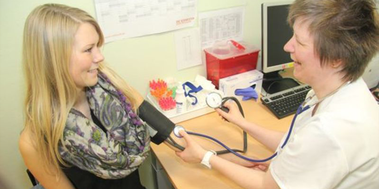 Eine Frau misst den Blutdruck einer anderen Frau in einem Büro. Beide lächeln.
