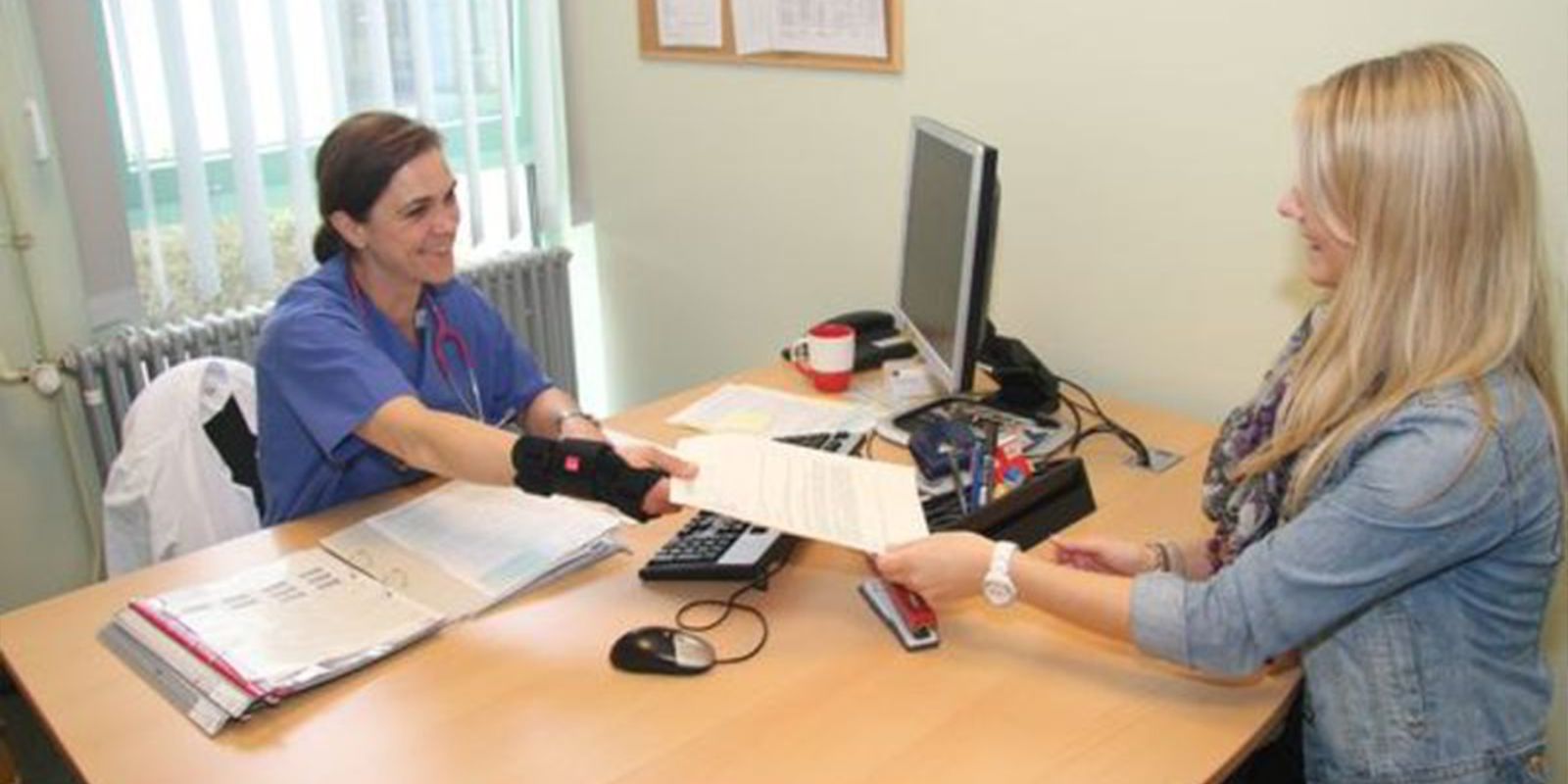 Eine Frau in einem Büro reicht einer anderen Frau hinter dem Schreibtisch ein Dokument. 