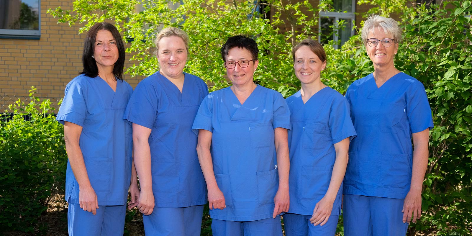 Fünf Menschen in blauer medizinischer Kleidung stehen draußen vor grünem Hintergrund.