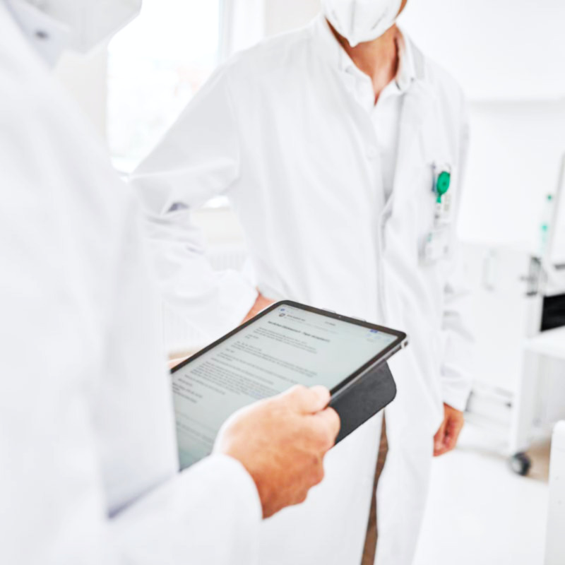 . Zwei Personen in weißen Kitteln und Masken, eine hält ein Tablet mit Text.