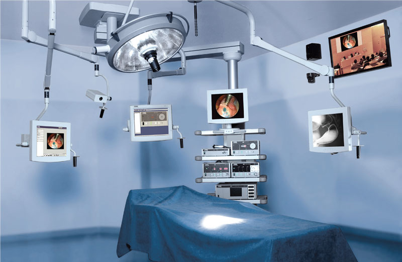 Ein Operationssaal mit blauem Tisch, mehreren Bildschirmen, medizinischen Geräten und Beleuchtung über Kopf.