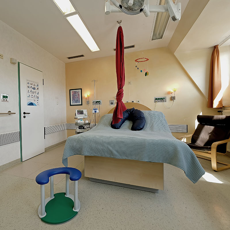 Ein Krankenhauszimmer mit einem Bett, Geräten, einem roten Tuch von der Decke und einem Sitzhocker.