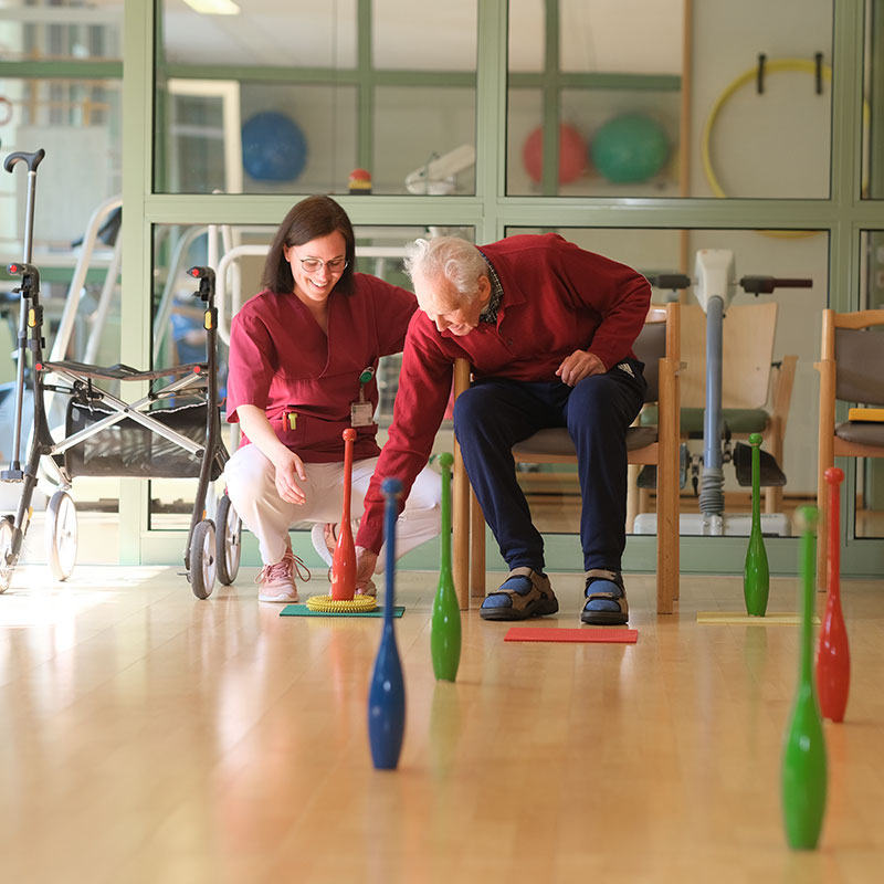 Eine Frau und ein älterer Mann üben mit bunten Kegeln in einem Raum mit Reha-Ausrüstung.