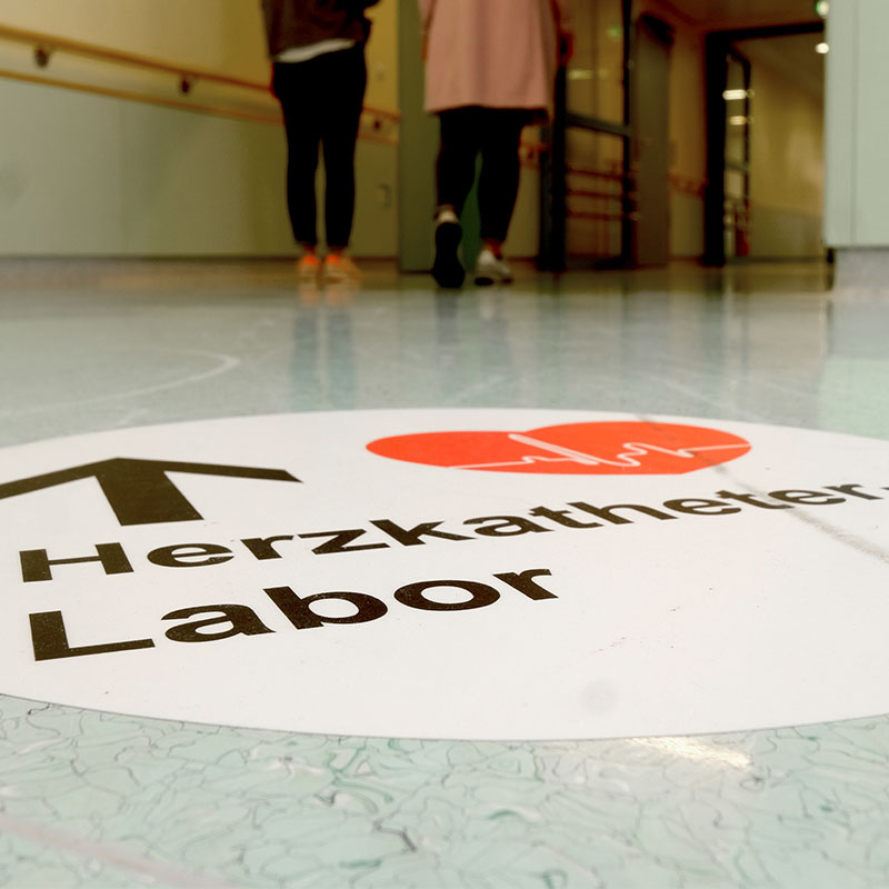 Bodenaufkleber mit Pfeil zum Herzkatheterlabor. Im Hintergrund gehen Menschen.