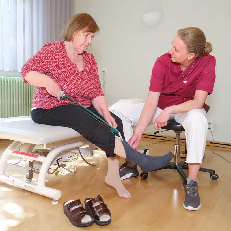 Eine ältere Frau zieht Socken an, unterstützt von einer Pflegekraft.