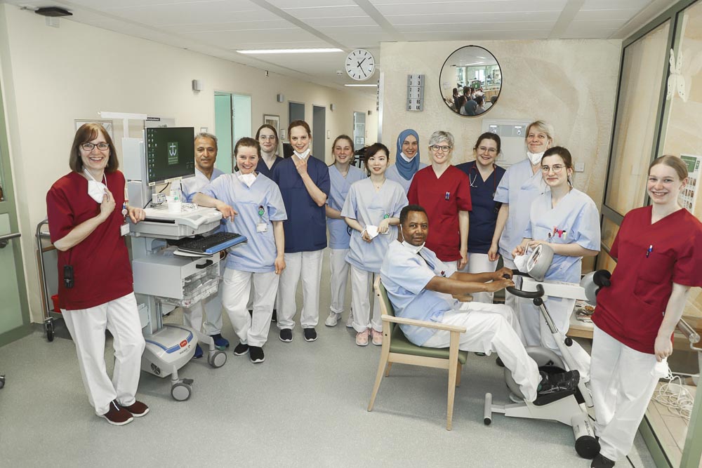 Eine Gruppe medizinischer Fachkräfte in Krankenhauskleidung posiert mit einem Fitnessgerät in einem Klinikraum.