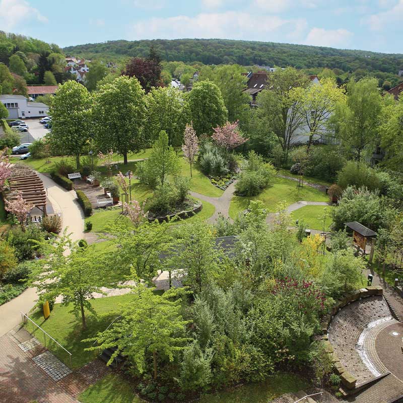 Ein grüner Park mit Wegen, Bäumen und Bänken, umgeben von Hügeln im Hintergrund.