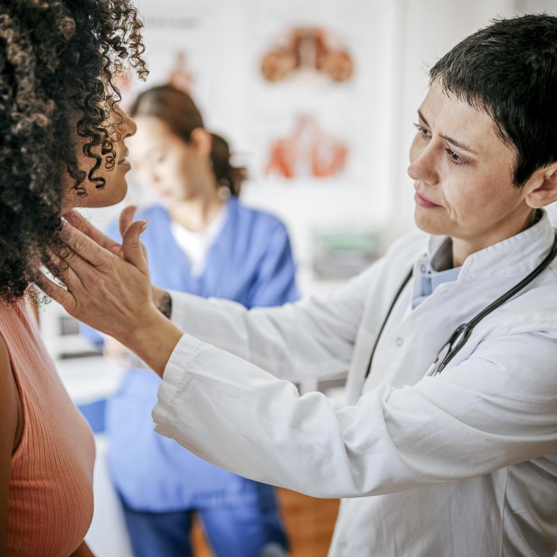 Ein Arzt untersucht eine Patientin in einer Praxis, während eine weitere Person im Hintergrund arbeitet.