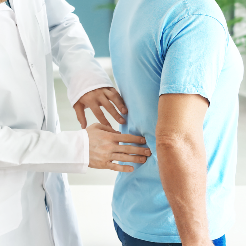 . Ein Arzt in weißem Kittel untersucht den unteren Rücken eines Mannes im blauen T-Shirt.