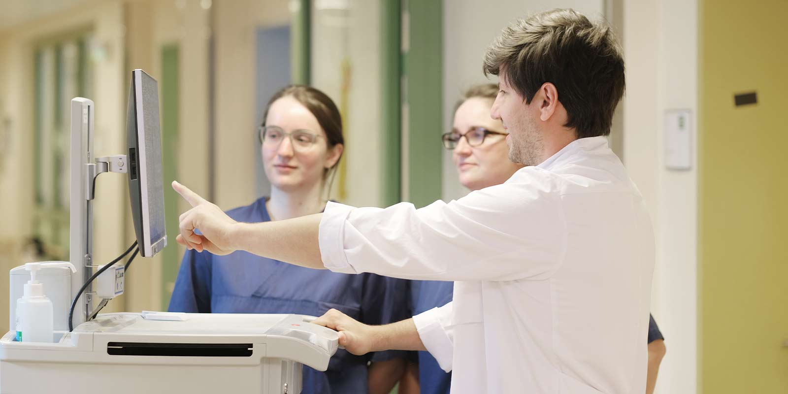 Ein Arzt zeigt den beiden Ärztinnen auf einem Monitor etwas in einem Krankenhausflur.