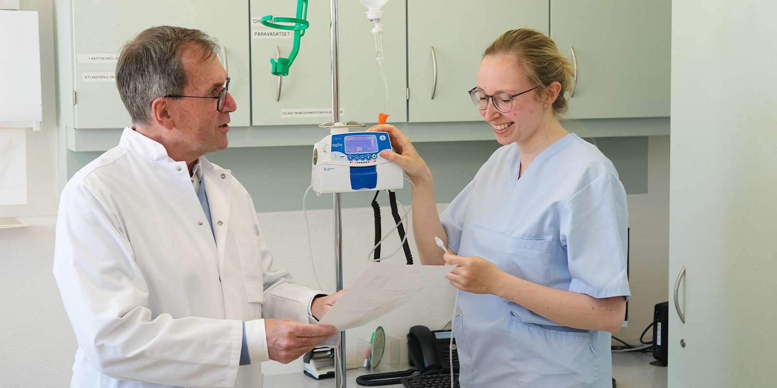 Ein Arzt und eine Krankenschwester lächeln und besprechen ein Dokument neben einem Infusionsgerät.