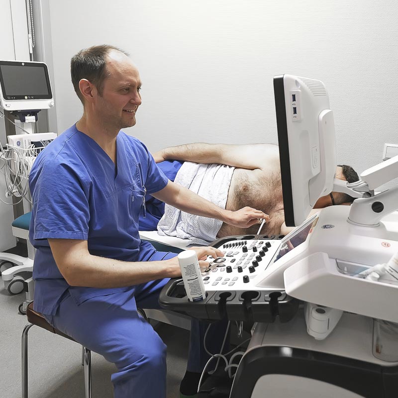 Ein Arzt in blauer Kleidung führt einen Ultraschall bei einem liegenden Patienten durch.
