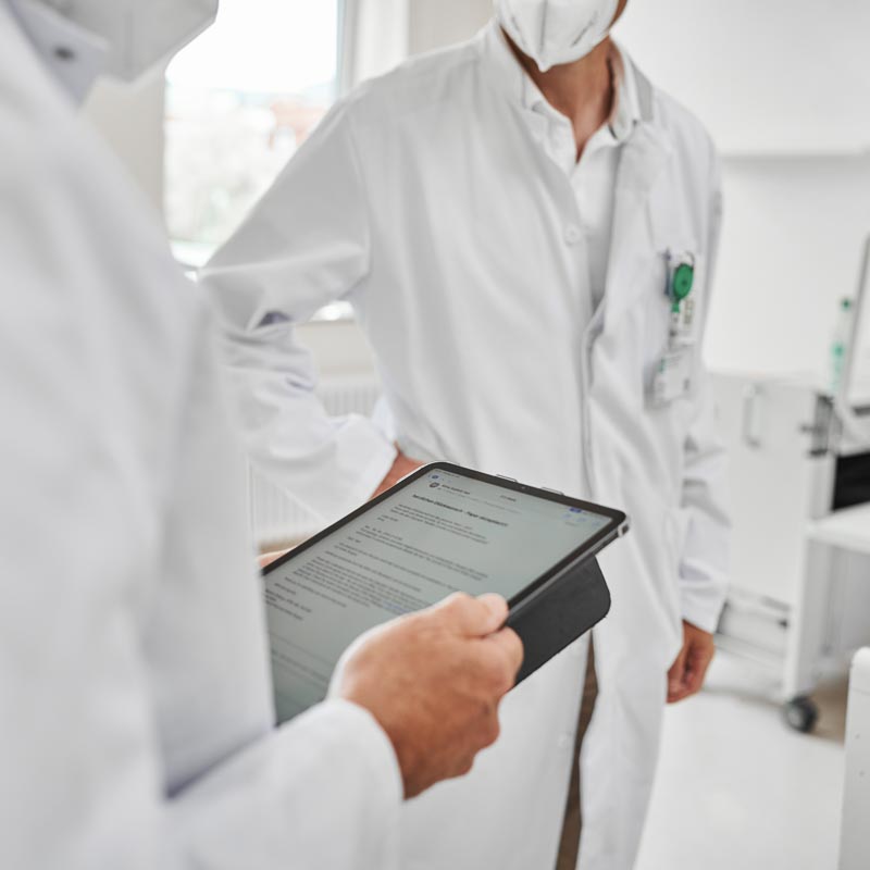 Zwei Personen in weißen Kitteln, eine hält ein Tablet mit Text. Sie scheinen in einem medizinischen Umfeld zu sein.