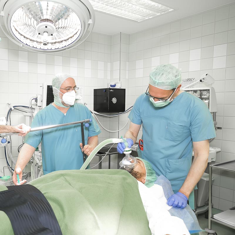 Ärzte im Operationssaal, die einen Patienten intubieren. Medizinische Ausstattung im Hintergrund.
