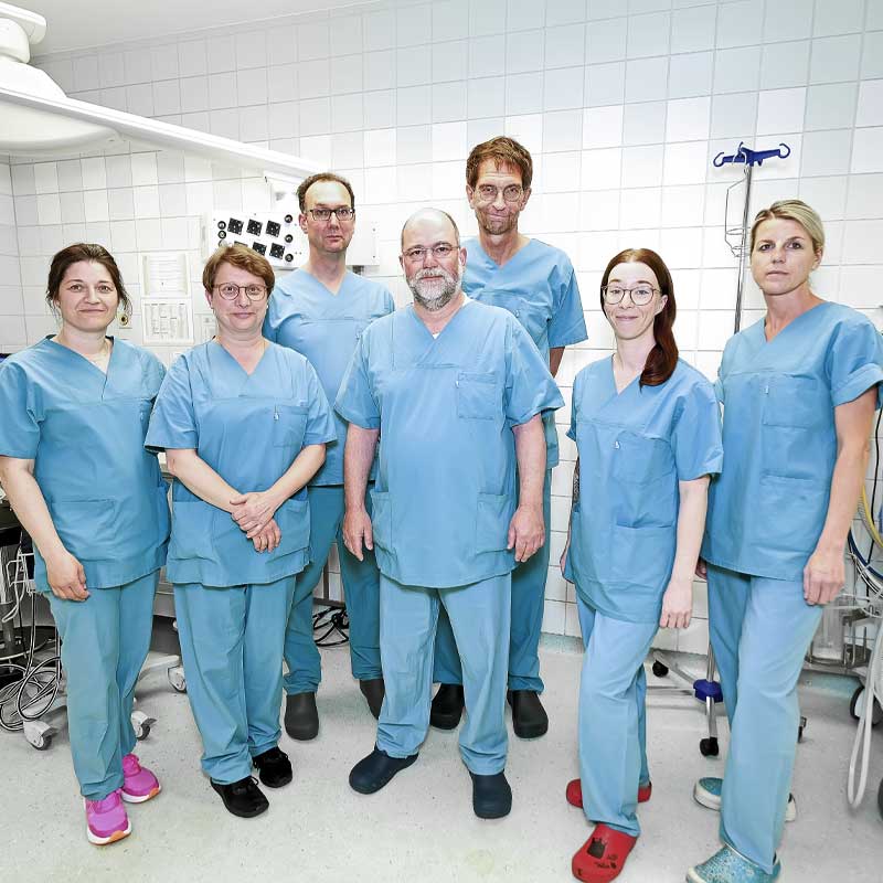 Eine Gruppe von sieben Personen in blauer Krankenhauskleidung posiert in einem medizinischen Raum.